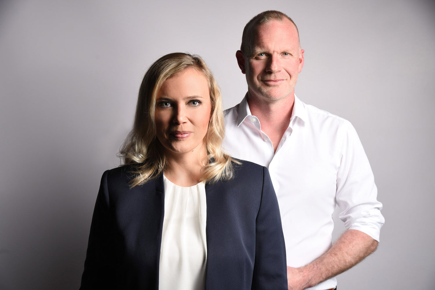 Zwei Personen, eine Frau im Vordergrund mit blonderen Haaren und dunklem Blazer und ein Mann im Hintergrund mit weißem Hemd, vor grauem Hintergrund.